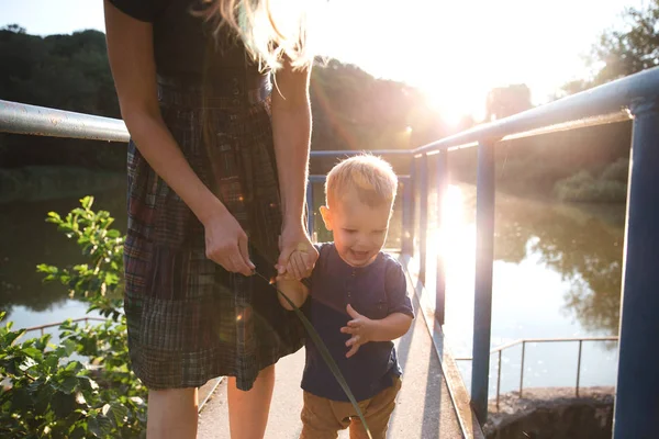 Front view mother and baby son hugging on the bridge on lake in summer evening time — Stock Photo, Image