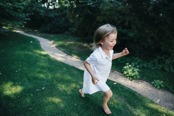 Vacker liten baby flicka, har glad kul leende ansikte, Blonda vackra hår, spelar i en vit klänning, barn porträtt. Barn familj. Fantastisk panoramautsikt. — Stockfoto