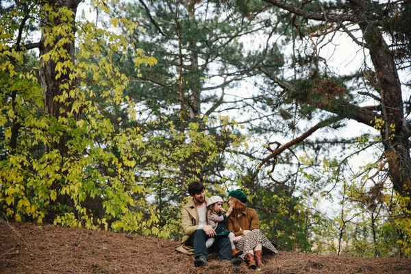 Junger Vater, Mutter und kleine Tochter in Baskenmütze und Mantel auf dem Boden im herbstlichen Wald. — Stockfoto