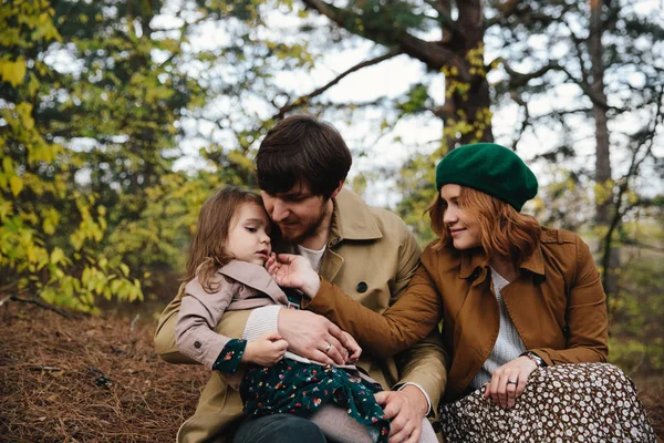 Jovem pai, mãe e filha pequena menina em uma boina e um casaco sentado no chão na floresta de outono . — Fotografia de Stock