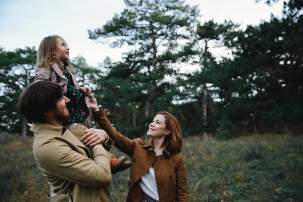 Ung far, mor och lite småbarn dotter flicka i en basker och en kappa försiktigt titta på varandra i höst skogen. — Stockfoto