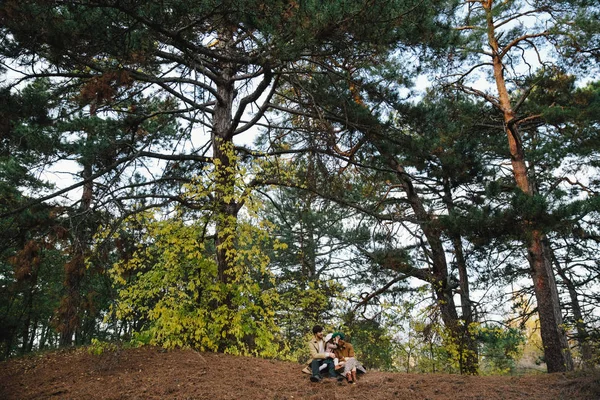 Young father, mother and little toddler daughter girl in a beret and a coat sitting on the ground in the autumn forest. — Stock Photo, Image