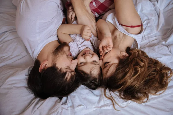 Family portrait of mother, father and daughter in white clothes having fun on the bed — Stock Photo, Image