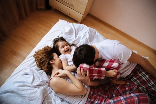 Family portrait of mother, father and daughter in white clothes having fun on the bed