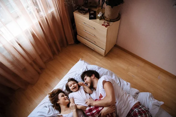 Family portrait of mother, father and daughter in white clothes having fun on the bed