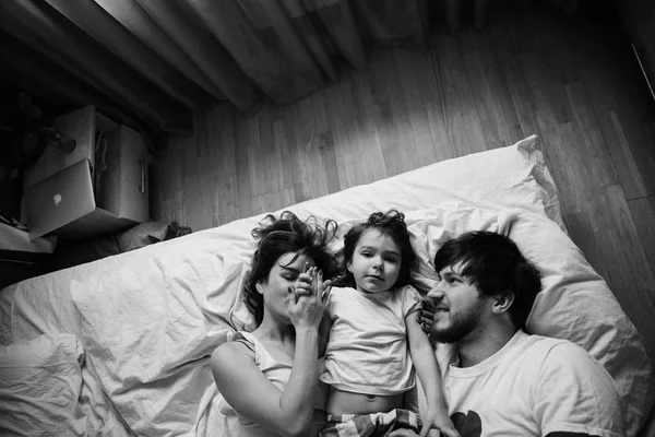 Black and white Family portrait of mother, father and daughter in white clothes having fun on the bed