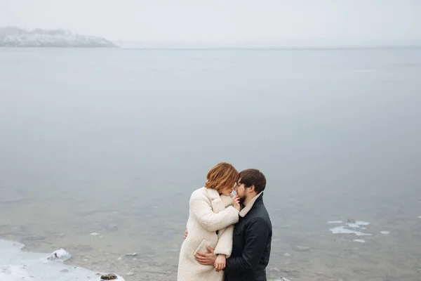 A couple in coats hugging on the banks of a frozen river — Stock Photo, Image
