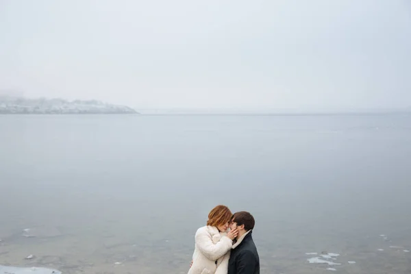 A couple in coats hugging on the banks of a frozen river — Stock Photo, Image