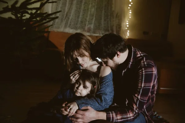Retrato de família de mãe, pai e filha em roupas de Natal abraçando no chão na escuridão e árvore de Natal perto deles — Fotografia de Stock