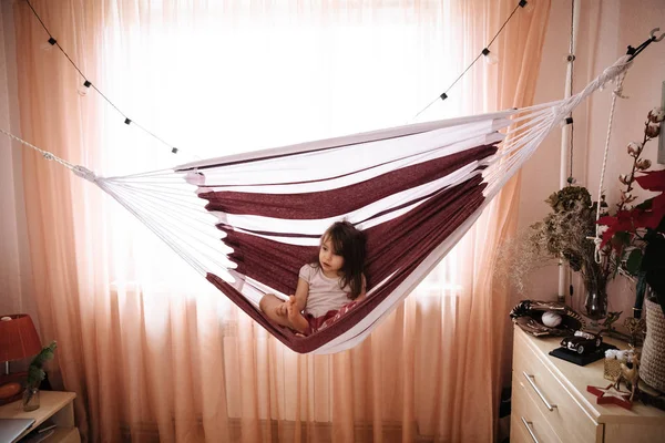 Full length portrait of pretty little girl with long hair in Christmas trouses and white T-shirt sitting comfortably in hammock and smiling at camera.
