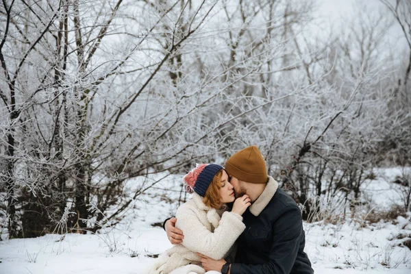 Mujer feliz y hombre con abrigos y sombreros sentados y besándose en el fondo de nieve de invierno. Concepto de pareja feliz y emociones positivas — Foto de Stock