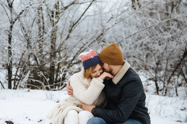 Glückliche Frau und Mann in Mänteln und Hüten sitzen und umarmen sich im winterlichen Schneehintergrund. Konzept des glücklichen Paares und positiver Emotionen — Stockfoto