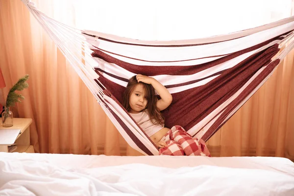 Full längd porträtt av söt liten flicka med långt hår i julen byxor och vit T-shirt sitter bekvämt i hängmattan och leende på kameran. — Stockfoto