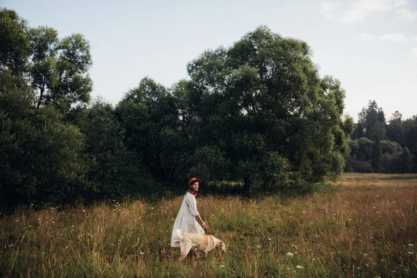 Beautiful young woman relaxed and carefree enjoying a summer sunset with her lovely dog on the straw field background — Stock Photo, Image