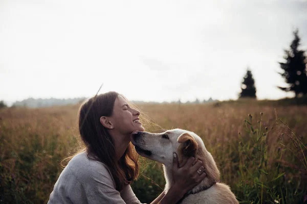 Hermosa mujer joven relajada y despreocupada disfrutando de una puesta de sol de verano con su hermoso perro en el fondo del campo de paja —  Fotos de Stock