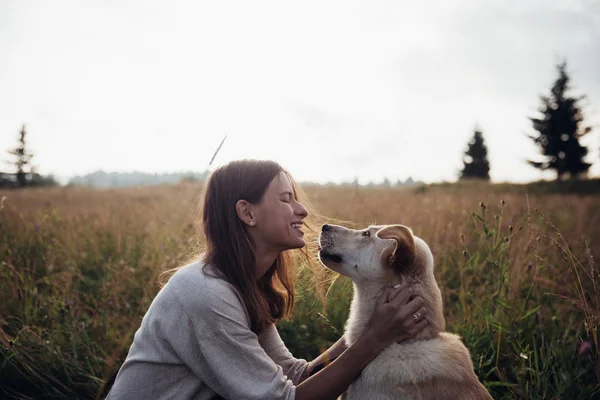 Hermosa mujer joven que se divierte al atardecer de verano con su hermoso perro en el fondo del campo de paja — Foto de Stock
