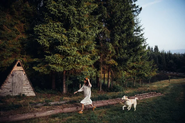 Woman pretty wear amazing white dress and hat are walking with a dog on forest and wooden wigwam background . Perfect place for ceremony. Stock Image