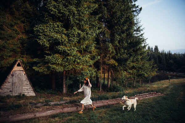 Woman pretty wear amazing white dress and hat are walking with a dog on forest and wooden wigwam background . Perfect place for ceremony. — Stock Photo, Image
