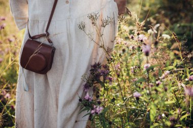 Gün batımı ışığında çayır kahverengi çanta ile keten elbise şık kız. Boho kadın rahatlatıcı ve yaz tarlasında kır çiçekleri toplama. Atmosferik kırsal an