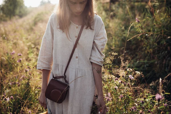 Stylisches Mädchen im Leinenkleid mit brauner Tasche auf der Wiese im Sonnenuntergangslicht. Boho Frau entspannen und sammeln Wildblumen im Sommer Feld. Stimmungsvoller Moment auf dem Land — Stockfoto