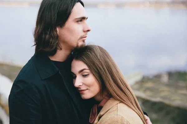 Giovane coppia romantica in cappotti abbracciando e godendo paesaggio autunnale sullo sfondo del fiume — Foto Stock