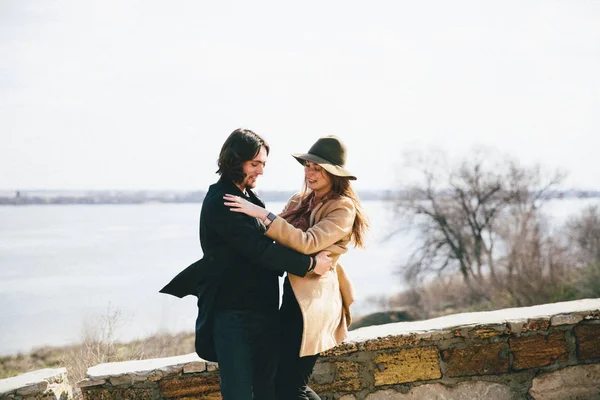 Young romantic couple in coats hugging and enjoying autumn landscape on the river background — Stockfoto