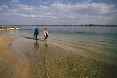 Sıcak paltolu genç çift nehir kıyısında eğleniyor. Nehirde yürümek