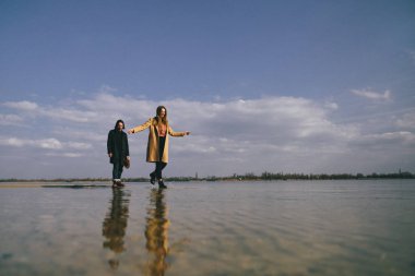 Sıcak paltolu genç çift nehir kıyısında eğleniyor. Nehirde yürümek