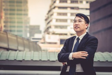 Zeki Asyalı iş adamı gülümseyerek kollarını göğsünün üzerine kavuşturur. Kollarını kavuşturup halefi düşünüyor.