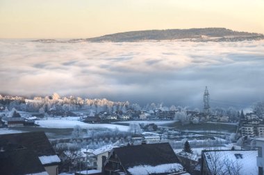 İsviçre 'de sabah manzarası, çatılar ve soğuk ağaçlarla vadiyi kaplayan dramatik sis denizi, yumuşak altın ışığın altında