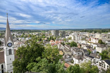 Uster, İsviçre 'nin panoramik manzarası, kilise kulesini saat, konut çatıları, modern binalar ve parçalı bulutlu gökyüzü altında yeşil çevreler ile gösteriyor.