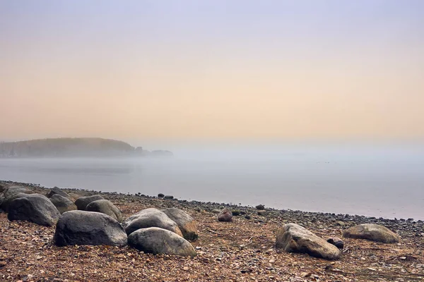 Lakeside shore and islands hidden behind thick fog. Stones on the coast; sand and pebble beach. Early morning; misty landscape.