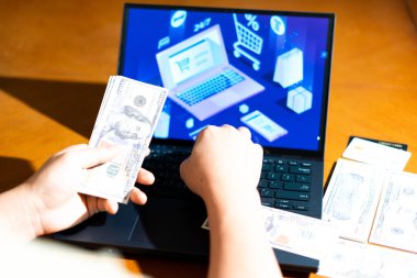 Bilgisayarın önünde ABD Doları banknotları tutan kişi, e-ticaret kontrol ekranına bakarken, finans blogları, dijital alışveriş temaları ve para yönetimi görselleri için.
