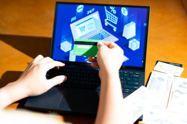 Masada USD banknotlarla online alışveriş yaparken kredi kartı tutan bir tüketicinin omuz üstü görüntüsü. Bir dijital çıkış işlemi sırasında nakit ve kart arasında satın alma kararı.