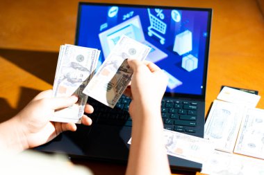 Bilgisayarın önünde ABD Doları banknotları tutan kişi, e-ticaret kontrol ekranına bakarken, finans blogları, dijital alışveriş temaları ve para yönetimi görselleri için.