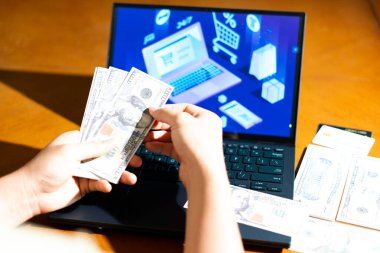 Bilgisayarın önünde ABD Doları banknotları tutan kişi, e-ticaret kontrol ekranına bakarken, finans blogları, dijital alışveriş temaları ve para yönetimi görselleri için.