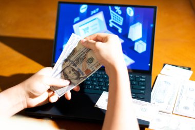 Bilgisayarın önünde ABD Doları banknotları tutan kişi, e-ticaret kontrol ekranına bakarken, finans blogları, dijital alışveriş temaları ve para yönetimi görselleri için.
