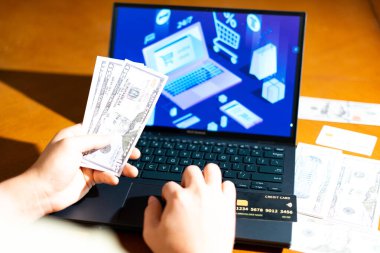 Bilgisayarın önünde ABD Doları banknotları tutan kişi, e-ticaret kontrol ekranına bakarken, finans blogları, dijital alışveriş temaları ve para yönetimi görselleri için.