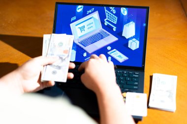 Bilgisayarın önünde elinde ABD Doları banknotları tutan birinin e-ticaret sayfası olan bir fotoğrafı. Geleneksel nakit alışveriş. Ödeme seçeneği, finansal kapsama veya harcama alışkanlıkları.