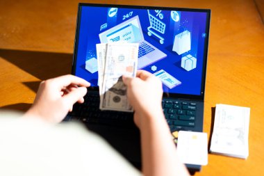 Bilgisayarın önünde elinde ABD Doları banknotları tutan birinin e-ticaret sayfası olan bir fotoğrafı. Geleneksel nakit alışveriş. Ödeme seçeneği, finansal kapsama veya harcama alışkanlıkları.