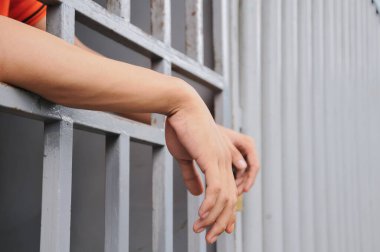Close-up, candid shot of a person's hands gripping the bars of a gray metal fence, with a building in a blurred background, representing detention or prison.