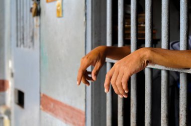 Close-up, candid shot of a person's hands gripping the bars of a gray metal fence, with a building in a blurred background, representing detention or prison.