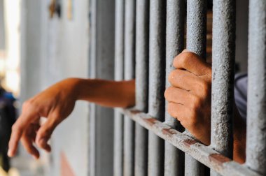 Close-up, candid shot of a person's hands gripping the bars of a gray metal fence, with a building in a blurred background, representing detention or prison.