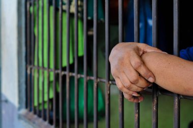 Close-up, candid shot of a person's hands gripping the bars of a gray metal fence, with a building in a blurred background, representing detention or prison.