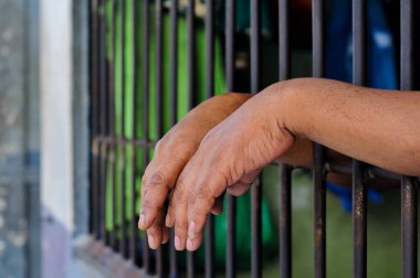 Close-up, candid shot of a person's hands gripping the bars of a gray metal fence, with a building in a blurred background, representing detention or prison.