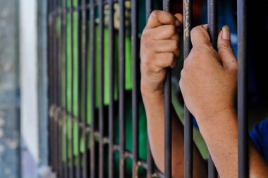 Close-up, candid shot of a person's hands gripping the bars of a gray metal fence, with a building in a blurred background, representing detention or prison.