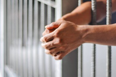 : A close-up of a person's hands gripping the cold, gray metal bars of a jail cell, with the background blurred, conveying a feeling of sadness, confinement, and longing for freedom.