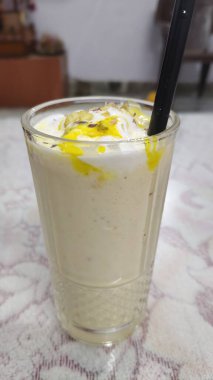 Kremalı, bej renkli, krem şantili ve sarı şerbetli bir içecekle dolu uzun bir bardak. Mango Milk shake. İçecekten siyah bir kamış uzanıyor ve üzerine fındık ya da çekirdek serpiştirilmiş..