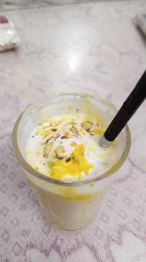 Bir bardak mango lassi, popüler bir Hint içeceği, resmedildi. Badem dilimleri (Prunus dulcis) ve safran iplikleri (Crocus sativus sativus) ile kaplı sarı renkli kremalı ve beyazdır.).