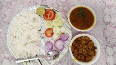 Bir tabak domates, salatalık, limon ve kırmızı soğan dilimlerinin yanında beyaz pirinç içerir. Pirincin yanında gümüş bir kaşık yatıyor..